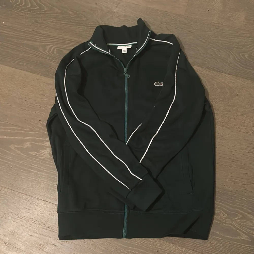 Säljer en snygg svart tröja med dragkedja från Lacoste. Den har vita detaljer längs ärmarna och det klassiska krokodilmärket på bröstet. Perfekt för en casual look eller träning. Tröjan är i mycket bra skick och redo för nya äventyr!. Jackor.