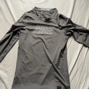 Grå långärmad träningströja från Nike - Säljer en grå långärmad träningströja från Nike Pro. Perfekt för träning med sitt bekväma och stretchiga material. Tröjan har en stilren design med Nike-loggan på bröstet. Passar bra för både inomhus- och utomhusaktiviteter.