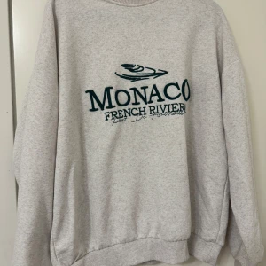 Grå sweatshirt med Monaco-tryck - Säljer en grå sweatshirt med ett coolt Monaco French Riviera-tryck i mörkgrönt på framsidan. Tröjan är i bra skick och är i storlek: XS men är oversized i modellen. Den har långa ärmar och ribbade muddar vid ärmslut och nederkant. Perfekt för höst och vår!