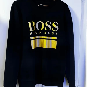 Svart tröja från Hugo Boss - Säljer en stilren svart tröja från Hugo Boss med ett stort guldtryck på framsidan. Tröjan har långa ärmar och är perfekt för både vardag och fest. Den är i mycket bra skick och passar perfekt till höst- och vintergarderoben.