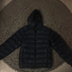 Marinblå dunjacka från Peak Performance - Säljer en marinblå dunjacka från Peak Performance i mycket bra skick. Jackan har en skön huva och dragkedja framtill. Perfekt för kalla vinterdagar och håller dig varm och bekväm. Passar både till vardags och utomhusaktiviteter.