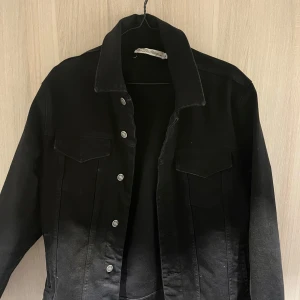 Svart denimjacka från The Blue Uniform - Säljer en stilren svart denimjacka från The Blue Uniform i storlek XL. Jackan har en klassisk design med knappar framtill och två bröstfickor. Perfekt för både vardag och festliga tillfällen. Passar bra till höst och vår!