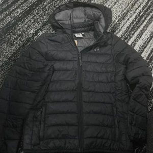 Svart pufferjacka från Hang Ten - Säljer en svart pufferjacka från Hang Ten i storlek 158/164. Jackan har en dragkedja framtill och är perfekt för kyliga dagar. Den är i bra skick och har en mysig huva för extra värme. Perfekt för både höst och vår.lite sönder men sydde den nära fickan. 