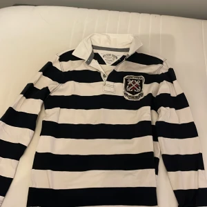 Randig pikétröja från Hilfiger Denim - Säljer en snygg randig pikétröja från Hilfiger Denim i svart och vit. Tröjan har långa ärmar och en cool broderad detalj på ryggen med texten 'New York'. Ralph lauren liknande! 