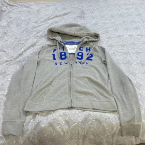 Grå hoodie från Abercrombie & Fitch - Säljer en snygg grå hoodie från Abercrombie & Fitch med blå text 'Fitch 1892 New York' på framsidan. Den har en dragkedja och är i bra skick. Perfekt för en avslappnad stil eller kyliga dagar. 🧢