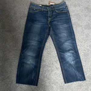 Blå jeans från Dondup - Säljer ett par snygga blå jeans oanvända från Dondup i modellen John. Storlek 28. De har en cool tvättad look med slitningar och kontrasterande gula sömmar. 