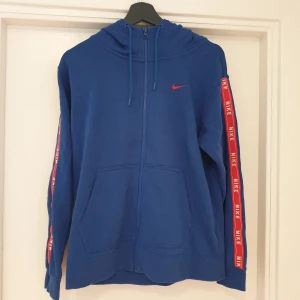 Blå hoodie från Nike - Säljer en snygg blå hoodie från Nike med dragkedja och huva. Den har röda detaljer med Nike-loggan längs ärmarna och en liten röd Swoosh på bröstet. Perfekt för en sportig look eller bara för att chilla. Superbekväm och i bra skick!