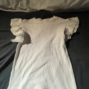 Vit ribbad topp från Gina Tricot - Säljer en superfin vit ribbad topp från Gina Tricot. Den har korta ärmar med volangdetaljer som ger en söt touch. Perfekt för både vardag och fest! Toppen är i bra skick och passar perfekt till jeans eller kjol.💓