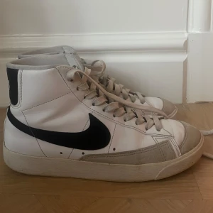 Vita Nike Blazers - Säljer ett par vita Nike sneakers i bra skick. De har en klassisk svart swoosh och snörning framtill. Skorna är höga och har en bekväm sula, perfekt för vardagsbruk. Litet skada på sulan men funkar perfekt. Perfekta för både vår och höst!