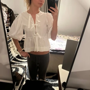 Vit blus från Ganni - Säljer en superfin vit blus från Ganni i mycket bra skick. Den har puffiga trekvartsärmar och söta knytband framtill. Perfekt för vår och sommar! Passar både till vardags och fest.
