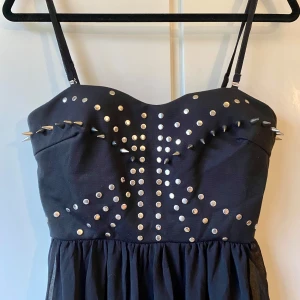 Svart klänning med nitar - Säljer en cool svart klänning med nitar på bröstet. Den har smala axelband och en asymmetrisk längd som är längre bak. Perfekt för en fest eller en utekväll! Klänningen är i bra skick och ger en edgy look.