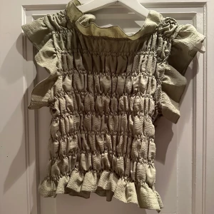 Grön topp med volanger - Säljer en superfin grön topp med volanger. Den har en smockad design som ger en cool textur och är perfekt för både vardag och fest. Toppen är ärmlös och har en söt volangdetalj vid ärmslut och nederkant. Saknas hänge till knappen baktill vid nacken. Man kan sy till ett nytt, eller bara inte knäppa (så har jag använt den och det har inte varit några problem/sett konstigt ut!) 