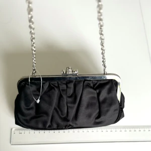 Svart kuvertväska med kedja - ALDO Svart handväska med en glittrig låsdetalj. Silverkedja.  Är perfekt för festliga tillfällen. Det får tex plats med en IPhone pro max & lite småsaker. Insidan har en liten ficka. Väskan är ny och redo för fest! Hämtas på Hägerstensåsen eller ök🖤