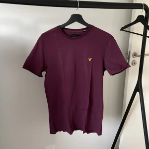 Vinröd t-shirt från Lyle & Scott - Säljer en snygg vinröd t-shirt från Lyle & Scott i storlek M. Den är kortärmad och har en broderad gul logga på bröstet. Perfekt för både vardag och fest!