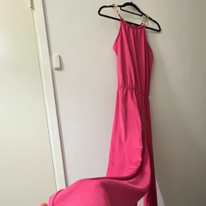 Rosa jumpsuit med kedjedetaljer - Säljer en superfin rosa jumpsuit med guldfärgade kedjor som axelband. Den är ärmlös och har en elastisk midja för en bekväm passform. Materialet känns mjukt och skönt mot huden.