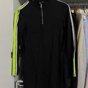 Svart klänning med neongula detaljer från Divided - Säljer en cool svart klänning från Divided med neongula och vita ränder längs ärmarna. Klänningen har en dragkedja framtill och är långärmad. Perfekt för en sportig look eller en avslappnad dag ute. Passar bra till sneakers eller boots!