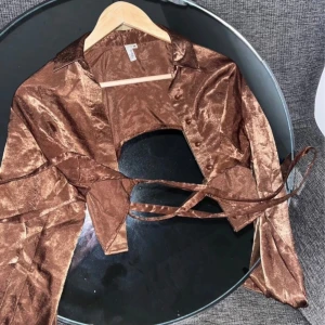 Croppad satinskjorta med öppen rygg samt snörning  - Endast använd en gång 🫶 Väldigt croppad i ryggen, snörning som man knyter hur man vill. Skimrigt skönt satin-material med knappar framtill samt vid handlederna.