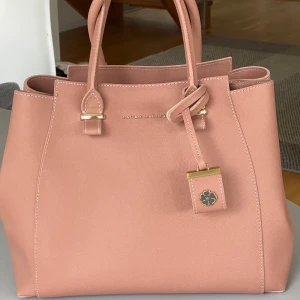 Laura Ashley handväska  - Laura Ashley väska i fin mörk laxrosa färg. Använd en gång och i nyskick. Inuti finns en stor ficka i mitten med blixtlås. Det finns ytterligare en ficka med blixtlås samt två mindre fickor. En avtagbar och justerbar axelrem som är oanvänd, se bild 3