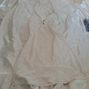 Vit klänning från H&M - Säljer en superfin vit klänning från H&M i storlek XS. Den är ärmlös och har en söt broderad design med små hål. Perfekt för sommardagar eller en festlig tillställning. Klänningen har en smickrande passform med knytband i ryggen. Materialet känns lätt och luftigt, vilket gör den idealisk för varma dagar.
