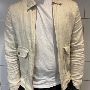 Beige linnejacka - Säljer en snygg beige jacka i linne. Den har en dragkedja framtill och två fickor med knappar. Perfekt för vår och sommar med sin lätta och luftiga känsla. Jackan har en normal passform och långa ärmar.