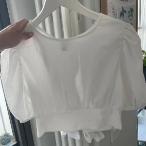 Vit kortärmad topp med knyt - Säljer en superfin vit topp från H&M. Den är kortärmad och har en söt knytning i ryggen som ger en extra detalj. Perfekt för sommardagar eller en avslappnad fest. Toppen är i ett lätt och luftigt material, vilket gör den bekväm att bära.