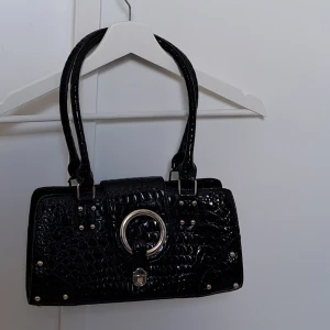 Svart shoulderbag - Medelstor axelväska i imitationsläder med krokodilmönster. Har ett spänne vid mitten som gör den perfekt att hänga bagcharms i. Fick ärva den av en kompis, så vet inte var den kommer ifrån.