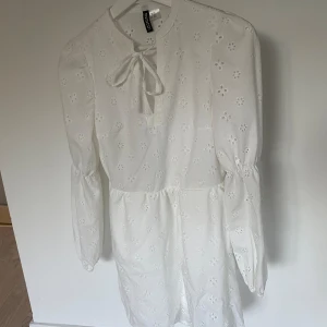 Vit klänning från H&M Divided - Säljer en superfin vit klänning från H&M Divided i storlek XXS. Klänningen har ett sött broderat blommönster och en knytning vid halsen. Den är långärmad och perfekt för vår och sommar. Materialet känns lätt och luftigt, vilket gör den idealisk för varmare dagar. Passar både till vardags och festligare tillfällen!