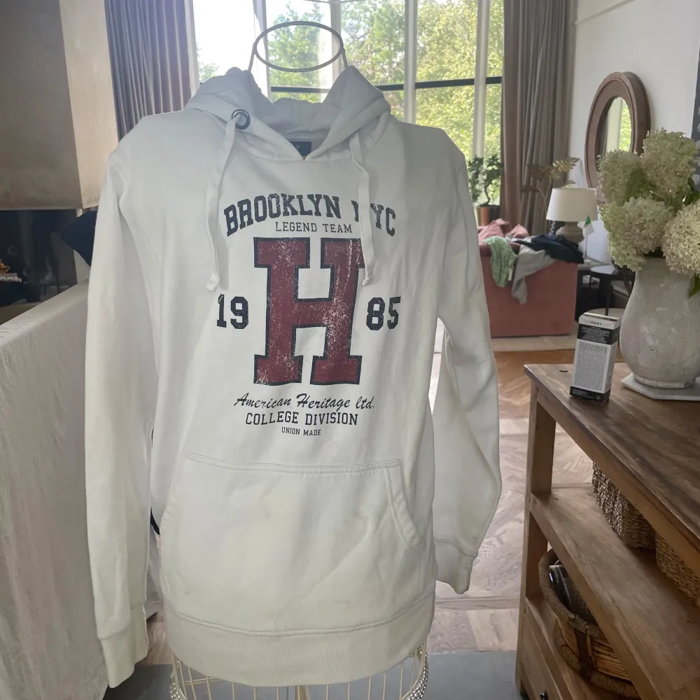 Säljer en snygg vit hoodie från Smog med tryck 'Brooklyn NYC Legend Team 1985' i vinrött och blått. Den har en stor bokstav 'H' på framsidan och texten 'American Heritage Club College Division'. Hoodien har en känguruficka och justerbar dragsko i huvan. Perfekt för en casual look!. Neuletakit & Villapaidat.