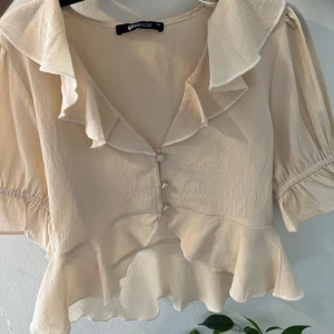 Beige blus från Gina Tricot - Säljer en superfin beige blus från Gina Tricot. Den har korta ärmar och en söt volangdetalj längs kragen och nederkanten. Perfekt för både vardag och fest!
