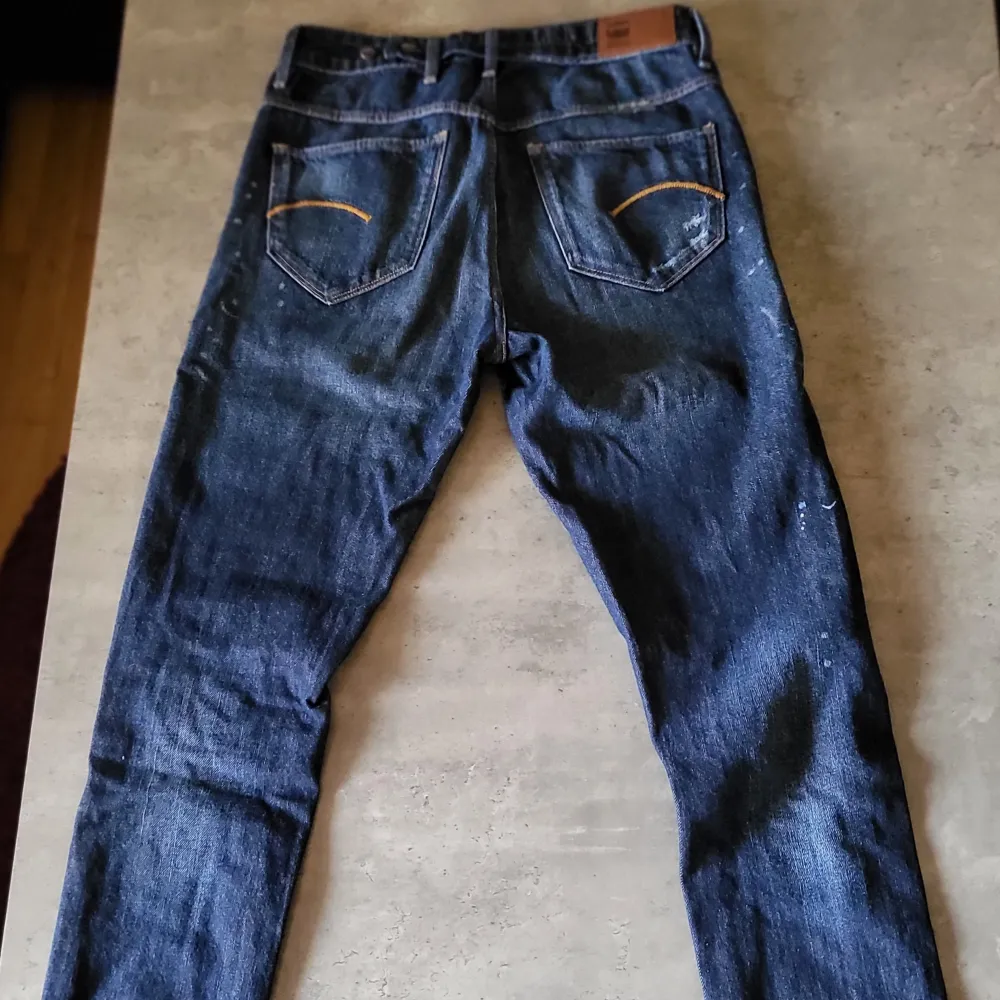 Säljer ett par snygga blå jeans från G-Star RAW. De har en cool slitning på knäna och en klassisk femficksdesign. Jeansen är i en straight fit och har en knappgylf. Perfekta för en avslappnad stil!. Jeans & Byxor.