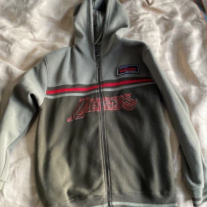 Grå hoodie från Anrtex - Säljer en grå hoodie från Anrtex i bra skick. Den har en dragkedja framtill och ett coolt Lakers-tryck på bröstet. Det finns också en patch på ärmen med en stjärna och text. Perfekt för höst och vinter, och den är superbekväm att ha på sig. Passar både till vardags och chillkvällar.