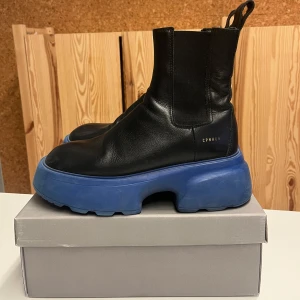 Svarta boots med blå sula  - Säljer ett par coola svarta boots i skinn från CPNHGN med en chunky blå sula. De har en stilren design med elastiska paneler på sidorna för enkel på- och avtagning. Perfekta för att ge din outfit en edgy touch! 🖤💙