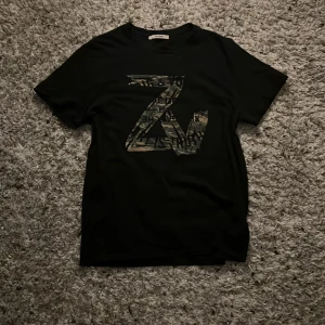 Svart t-shirt från Zadig & Voltaire - Snygg svart t-shirt från Zadig & Voltaire med ett stort ZV-tryck i camo-stil på framsidan. OBS Storlek XL men passar M-L. Perfekt för en casual look eller att styla upp med en cool jacka. T-shirten är i användt skick och därav de billiga priset. 