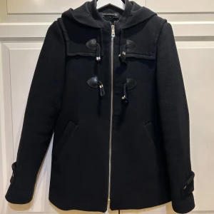 Svart kappa  - Säljer en stilren svart kappa från Zara i mycket bra skick. Den har en dragkedja framtill och snygga knappar som detaljer. Perfekt för vintern med sin varma design. Jackan har långa ärmar och en klassisk passform. Ett måste för garderoben! Köpte med en fuskpäls på men inget jag tyckte om och tog därför av den. Nypris 1200 men säljer för 499 eftersom pälsen inte medföljer. 