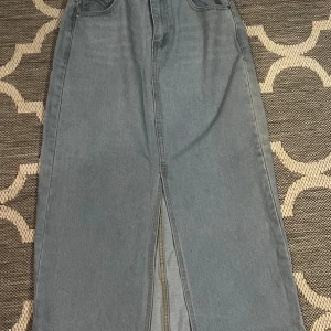 Lång jeanskjol med slits - Säljer en snygg lång jeanskjol i klassisk blå denim. Kjolen har en stilren design med en slits framtill som ger extra rörelsefrihet. Perfekt för både vardag och festliga tillfällen. Passar bra till en t-shirt eller en stickad tröja för en avslappnad look.