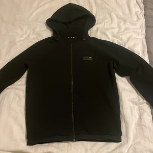 Svart EA7 hoodie - Säljer en svart hoodie från EA7 i mycket bra skick. Den har en dragkedja framtill och en bekväm huva. Perfekt för höst och vår, och funkar både till vardags och träning. Storleken är M. 