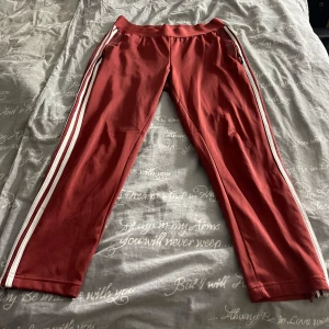 Röda träningsbyxor från Adidas - Snygga röda träningsbyxor från Adidas med vita ränder längs sidorna. Perfekta för träning eller en avslappnad dag. Byxorna har en elastisk midja för extra komfort. De är i bra skick och redo för nya äventyr!