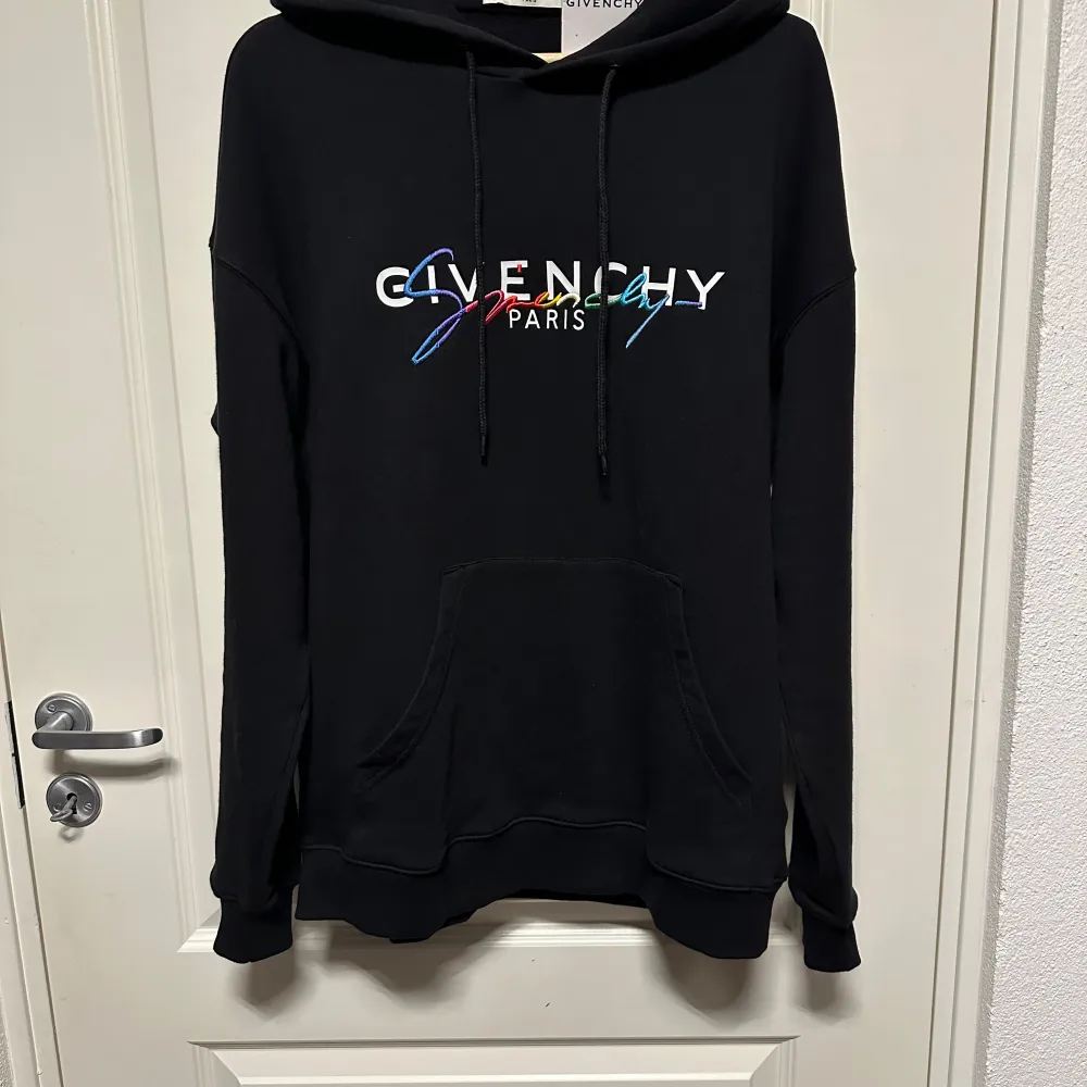 Säljer en snygg svart hoodie från Givenchy med färgglatt broderat logotyp på bröstet. Den har en klassisk känguruficka och justerbar dragsko i huvan. Perfekt för både vardag och chill. Helt ny med taggar kvar!. Hupparit & Collegepaidat.