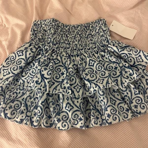 Mönstrad volangkjol - Superfin kortkjol med blått och vitt mönster. Kjolen har en elastisk midja och volanger som ger en söt och lekfull look. Perfekt för sommardagar eller en festlig kväll. Helt ny med tag kvar!