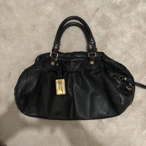 Svart handväska från Marc by Marc Jacobs - Snygg svart handväska i skinn från Marc by Marc Jacobs. Väskan har guldfärgade detaljer och en praktisk dragkedja. Insidan är fodrad med ett coolt mönster av märkets logga. Perfekt för både vardag och fest!