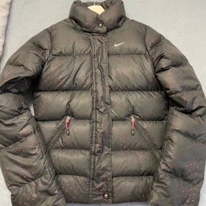 Nike jacka - Säljer en snygg, svart dunjacka från Nike med rosa prickar. Den är äkta såklart och nypriset är 1500kr. Den är perfekt för kalla vinterdagar eftersom den är så varm och puffig. Den har aldrig använts, så den är i nytt skick. Jackan är i storlek S men passar mig perfekt som har storlek Xs och sitter även perfekt på min mamma som har storlek M. Tveka inte med att skriva och priset kan sänkas i en snabb affär.