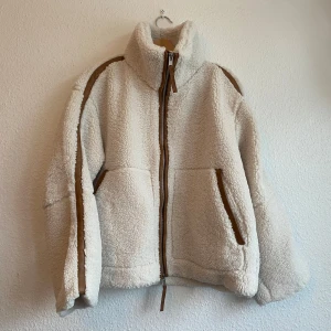 Ny cremevit teddyjacka med bruna detaljer storlek M från HM - Ny med tags. Supermysig vit pilejacka med bruna detaljer och dragkedja framtill. Perfekt för kyliga dagar med sin fluffiga och varma design. Jackan har en hög krage och två fickor. Jackan är ganska tjock då den är lätt vadderad.. storlek M (oversize)