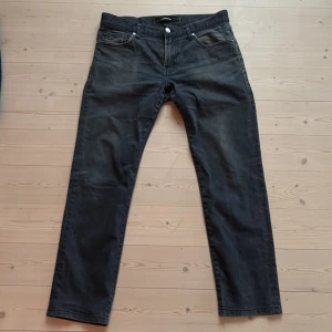 Svartgråa jeans från J.Lindeberg - Säljer ett par snygga gråsvarta jeans från J.Lindeberg i storlek 33/34 men är upplagda och snarare w 32/L 28. Innerbenlängd 71 cm, yttterbenlängd 92 cm, midja 43 cm (mätt rakt över linning). De har en klassisk femficksdesign och en knappgylf. Modell Jay Dressed, slim fit. 