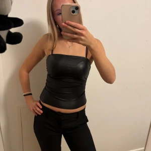 Svart tubtopp  - Snygg svart tubtopp i typ lackmaterial som ger en cool och stilren look. Perfekt för en utekväll eller fest. Toppen är tight och axelbandslös, vilket ger en smickrande passform. Passar perfekt till både jeans och kjol!