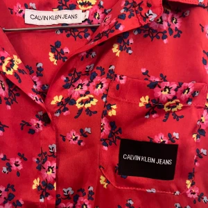 Röd blommig ärmlös topp från Calvin Klein Jeans - Säljer en superfin röd ärmlös topp från Calvin Klein Jeans med ett färgglatt blommönster i rosa, blått och gult. Perfekt för sommardagar eller en avslappnad vårlook. Toppen har en klassisk krage och en liten bröstficka med logga. Passar både till jeans och kjol!