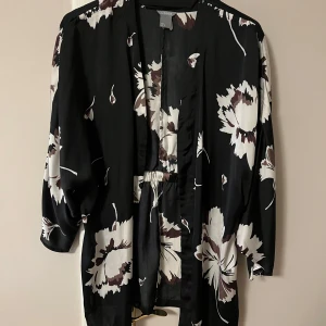 Svart blommig kimono - Säljer en superfin svart kimono från Lindex med stora vita och bruna blommor. Den har trekvartsärmar och ett knytband i midjan för en snygg passform. Perfekt för både vardag och fest! 🌸