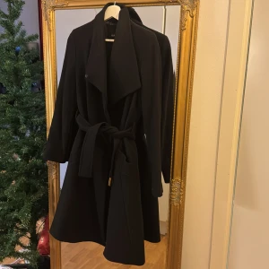 Svart kappa med bälte - Säljer en elegant svart kappa i mycket bra skick. Den har ett stilrent bälte i midjan och långa ärmar. Perfekt för höst och vinter, och funkar lika bra till vardags som till fest. Kappan har en klassisk design med en fin passform.