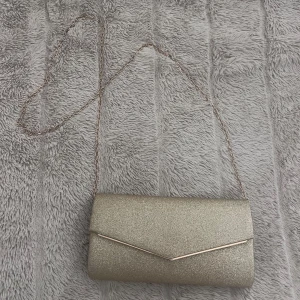 Glittrig guld kuvertväska med kedja - Säljer en superfin glittrig guld kuvertväska med kedja. Perfekt för fest, eller för att pimpa upp en vardaglig outfit. Den har en magnetisk knappstängning och en snygg design med en liten ficka inuti.  Väskan är använd 1 gång.