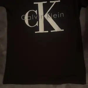 Säljer en stilren svart t-shirt från Calvin Klein med det ikoniska CK-loggan tryckt i vitt på framsidan. Perfekt för en casual look eller att klä upp med en snygg jacka. T-shirten är i bra skick och passar till alla tillfällen.