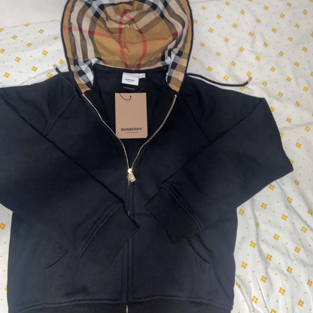 Säljer en snygg svart hoodie från Burberry med klassiskt rutigt mönster i luvan. Den har dragkedja framtill och är i mycket bra skick. Perfekt för höst och vår, och passar både till vardags och fest. Storleken är normal och materialet känns mjukt och bekvämt.. Hoodies.